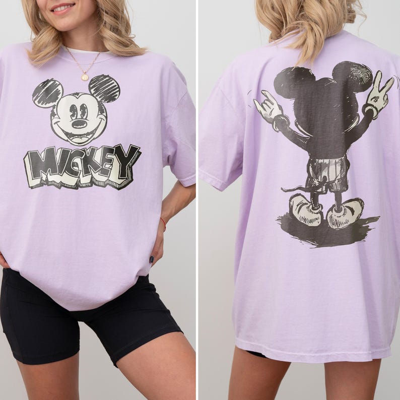 May include: A light purple short-sleeve t-shirt featuring a black and white sketch-style graphic of Mickey Mouse on the front with the text MICKEY below it, and a back graphic showing the character from behind with hands raised in a peace sign.