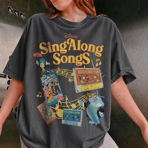 May include: A charcoal gray t-shirt featuring a graphic print with the text Disney Sing Along Songs in yellow. The design includes illustrations of Jiminy Cricket, a red Disneyland Fun cassette tape, a blue It's a Small World tape, and musical notes.
