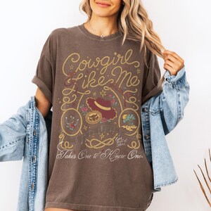 Pu&ograve; includere: Una t-shirt a maniche corte marrone scuro con una stampa grafica che riporta il testo Cowgirl like Me e Takes One to Know One. Il design include un cappello da cowboy, un ferro di cavallo, un bordo a corda, note musicali e piccoli fiori.