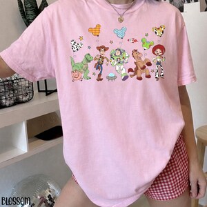 Puede incluir: Una camiseta de color rosa claro con un estampado gr&aacute;fico de los personajes de Toy Story, incluyendo a Woody, Buzz Lightyear, Jessie, Perdig&oacute;n, Rex y Hamm. El dise&ntilde;o incluye varios globos con forma de Mickey Mouse, estrellas y un cohete.
