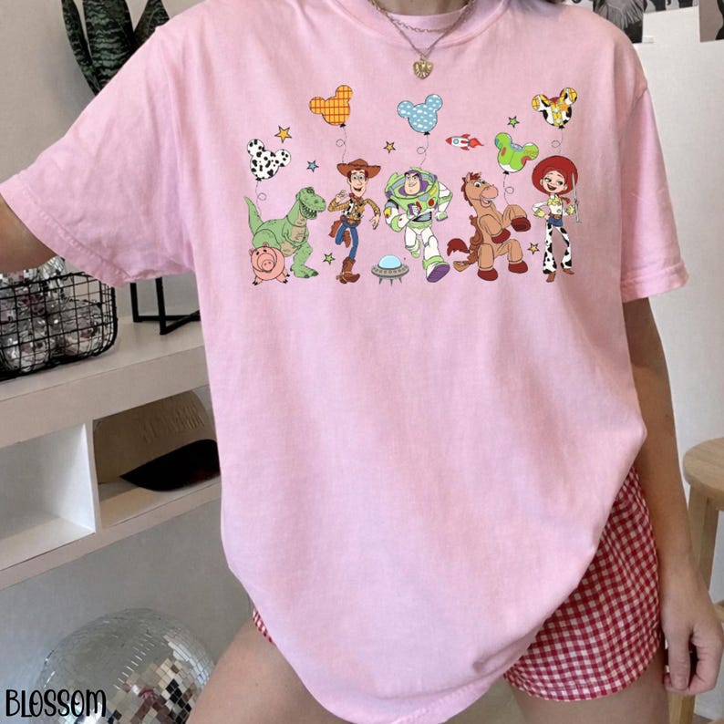 Puede incluir: Una camiseta de color rosa claro con un estampado gr&aacute;fico de los personajes de Toy Story, incluyendo a Woody, Buzz Lightyear, Jessie, Perdig&oacute;n, Rex y Hamm. El dise&ntilde;o incluye varios globos con forma de Mickey Mouse, estrellas y un cohete.