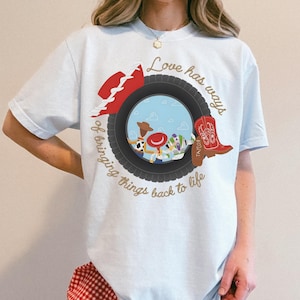 May include: A light blue t-shirt featuring a graphic of a black tire framing a scene with three cartoon characters. The design includes a red cowboy hat, a red cowboy boot, and the text "Love has ways of bringing things back to life" in gold.