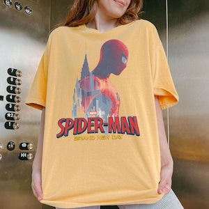 Op de afbeelding: Een geel t-shirt met korte mouwen en een afbeelding van Spider-Man in profiel tegen een stadssilhouet. Het ontwerp bevat het Marvel Studios-logo en de tekst "SPIDER-MAN BRAND NEW DAY" in dikke rode en blauwe letters op de voorzijde.