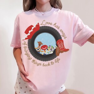 May include: A light pink t-shirt featuring a graphic of a black tire framing a scene with three cartoon characters. The design includes a red cowboy hat, a red cowboy boot with the name Taylor, and the text Love has ways of bringing things back to life.