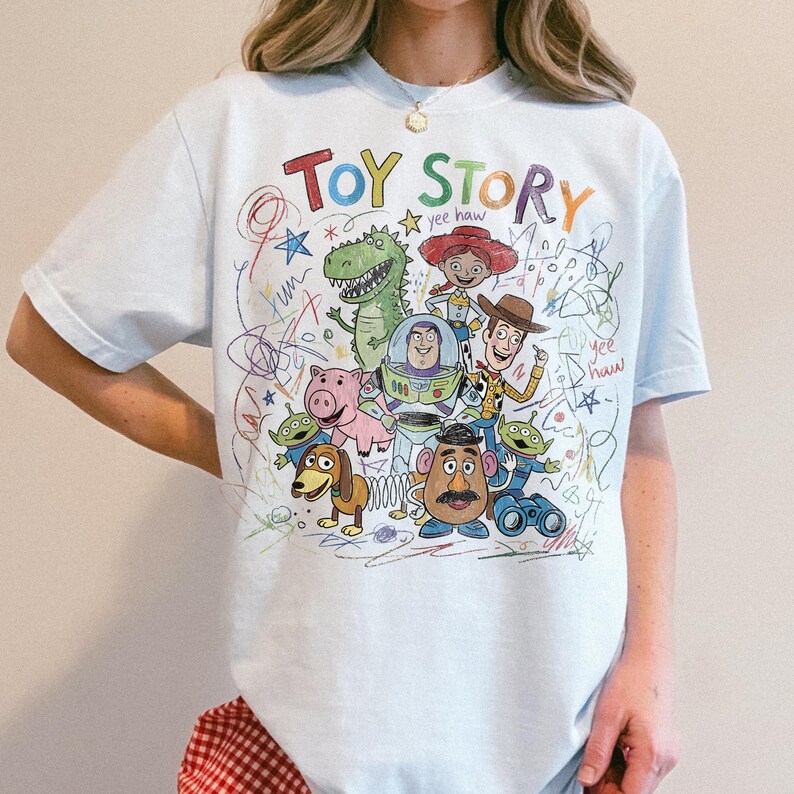 Peut inclure: T-shirt bleu clair avec un graphisme color&eacute; de style dessin&eacute; &agrave; la main repr&eacute;sentant les personnages de Toy Story. Le design comprend les mots "TOY STORY" dans une police ludique, ainsi que divers personnages.