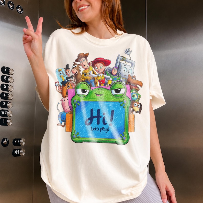 May include: An off-white t-shirt featuring a colorful graphic print of various animated characters from the Toy Story franchise. A central green frog-shaped frame displays the text Hi! Let's play! in a blue, crayon-like script on the front.