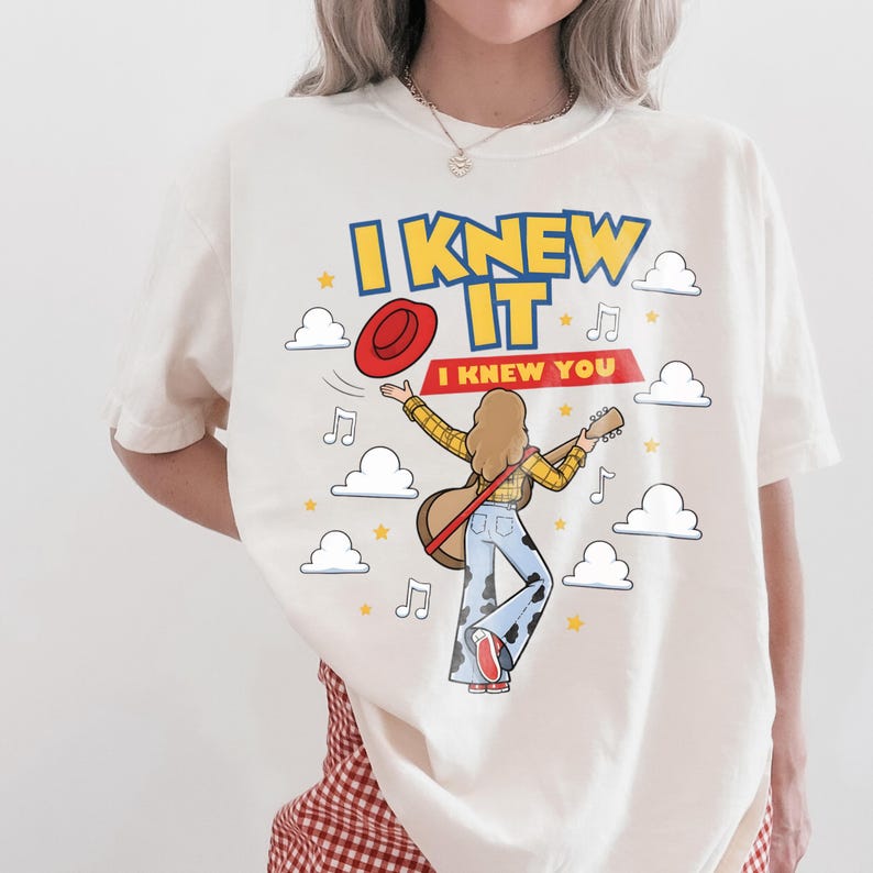 May include: An off-white t-shirt featuring a graphic of a person with long hair playing a guitar, wearing a yellow plaid shirt and cow-print bell-bottom jeans. The text reads I KNEW IT and I KNEW YOU, surrounded by red hat, clouds, and music notes.