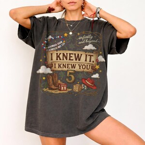 May include: A charcoal gray short-sleeve t-shirt featuring a graphic print with the text "I KNEW IT, I KNEW YOU", "infinity and beyond", and "YOU'VE GOT A FRIEND IN ME". The design includes a cowboy hat, boots, blocks, and stars.