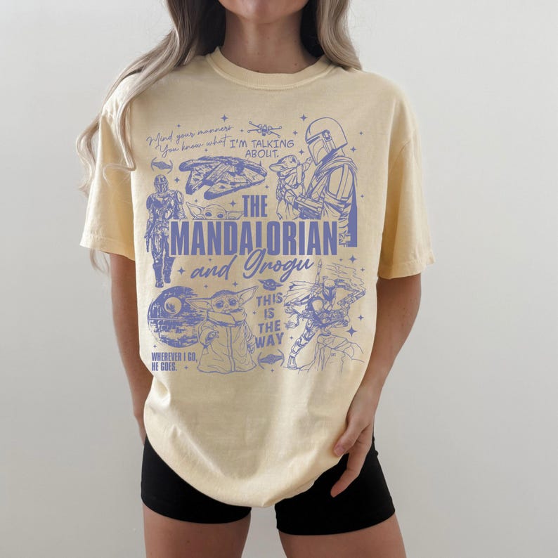 May include: A pale yellow short-sleeve t-shirt featuring a blue graphic print of The Mandalorian and Grogu. The design includes various character illustrations, the text "THE MANDALORIAN and Grogu", "THIS IS THE WAY", and "WHEREVER I GO, HE GOES."