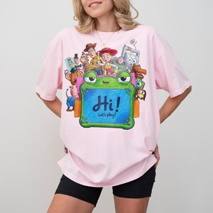May include: A light pink t-shirt featuring a graphic print of various animated characters from Toy Story surrounding a green frog-shaped chalkboard. The chalkboard has the text "Hi! Let's play!" written in blue script on its center surface.
