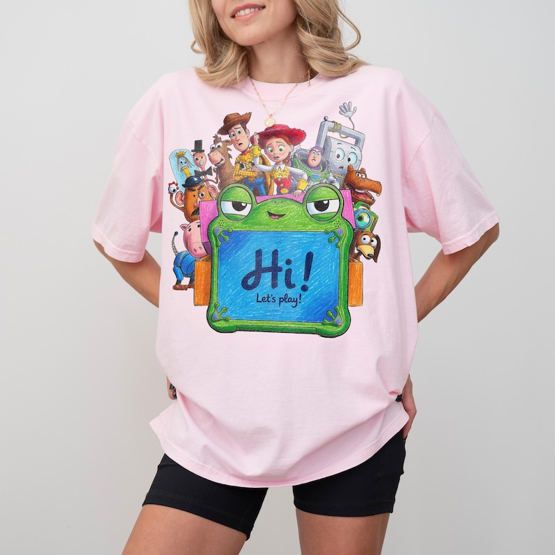 May include: A light pink t-shirt featuring a graphic print of various animated characters from Toy Story surrounding a green frog-shaped chalkboard. The chalkboard has the text "Hi! Let's play!" written in blue script on its center surface.