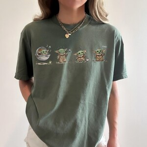 May include: An olive green short-sleeve t-shirt featuring a graphic print of a small green creature in four different poses. The illustrations show the character in a floating pod, using the force, sitting with a cup, and waving with the text "coo!"