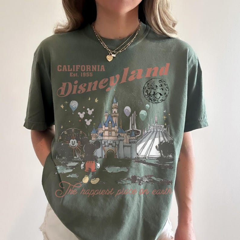 May include: Olive green t-shirt featuring a Disneyland graphic. The design includes the text "California Disneyland Est. 1955" and an illustration of the castle, Mickey Mouse, and other park elements. The phrase "The happiest place on earth" is also included.