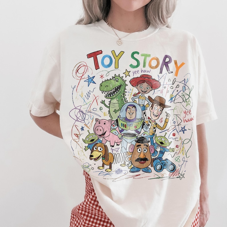 Peut inclure: T-shirt cr&egrave;me avec un dessin color&eacute; des personnages de Toy Story, dont Buzz l'&Eacute;clair et Woody. Les mots "TOY STORY" sont &eacute;crits dans une police multicolore ludique au-dessus des personnages. Le design a une esth&eacute;tique dessin&eacute;e &agrave; la main.