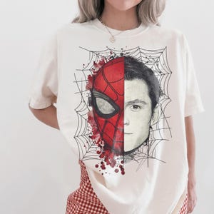 May include: An off-white short-sleeve t-shirt featuring a graphic print of a face split down the middle. One side shows a red and black Spider-Man mask with web patterns and red paint splatters, while the other side shows a realistic human face.
