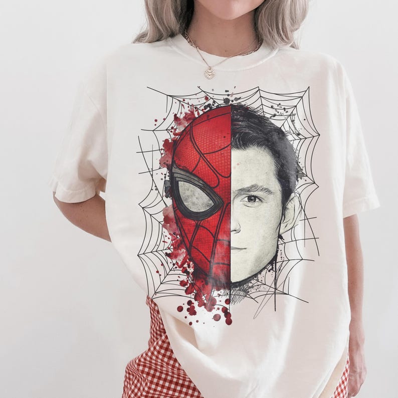 May include: An off-white short-sleeve t-shirt featuring a graphic print of a face split down the middle. One side shows a red and black Spider-Man mask with web patterns and red paint splatters, while the other side shows a realistic human face.