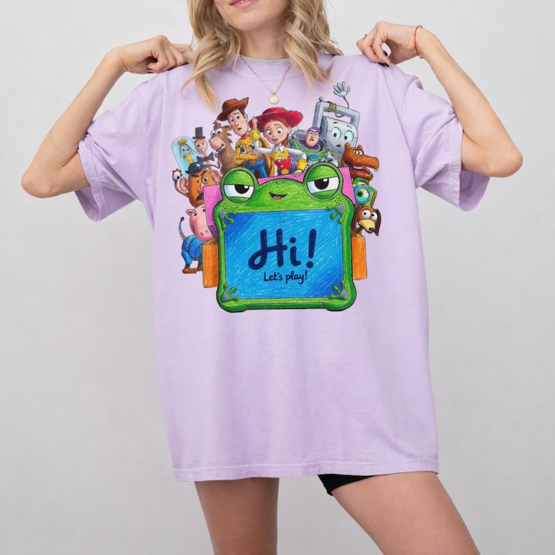 May include: A light purple t-shirt featuring a colorful graphic of various animated characters from the Toy Story franchise. In the center, a green frog-shaped frame contains the blue text "Hi! Let's play!" surrounded by characters.