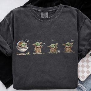 May include: A charcoal gray crew neck t-shirt featuring a graphic print of a small green creature in four different poses. The illustrations show the character in a floating pod, standing, sitting with a question mark, and waving with the text coo.