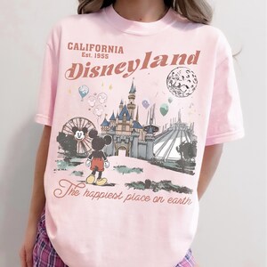 May include: Pink t-shirt featuring a Disneyland graphic. The design includes a castle, Mickey Mouse, a Ferris wheel, and the text "California Disneyland Est. 1955" and "The happiest place on earth."