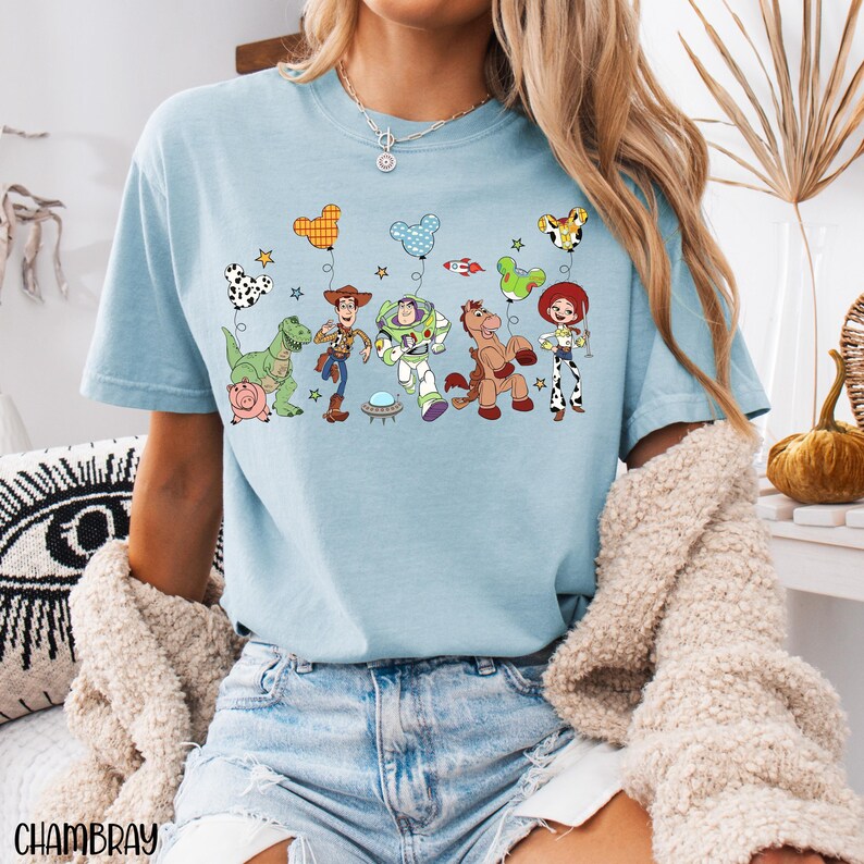 Puede incluir: Una camiseta azul claro con un estampado gr&aacute;fico de personajes de Toy Story, incluyendo a Woody, Buzz Lightyear, Jessie, Rex, Perdig&oacute;n y Hamm. Los personajes aparecen con globos de colores con forma de Mickey Mouse, estrellas y un cohete.