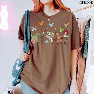 Puede incluir: Una camiseta marr&oacute;n de manga corta con un estampado gr&aacute;fico de varios personajes animados, incluyendo un vaquero, un guardi&aacute;n espacial, una vaquera, un dinosaurio, un cerdo y un caballo, junto a globos flotantes y el texto "espresso".