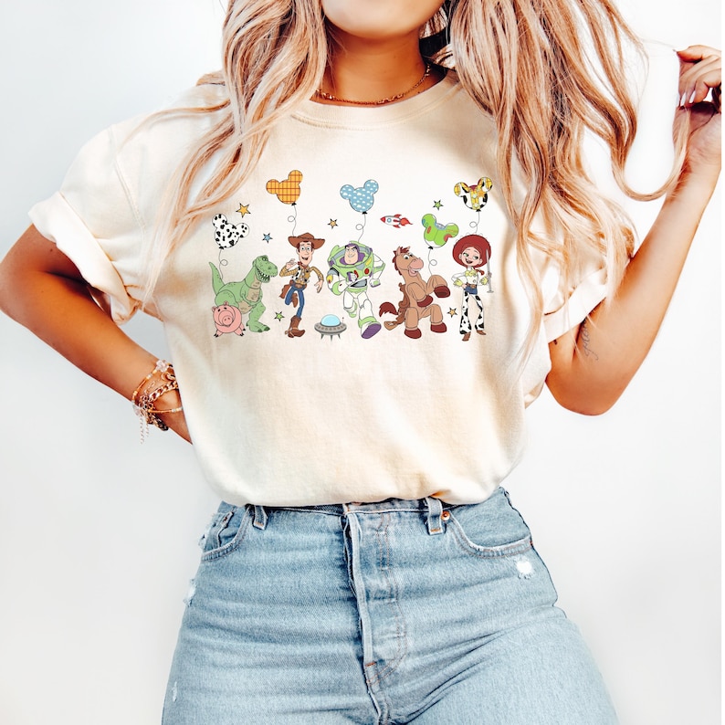 Puede incluir: Una camiseta de manga corta color crema con un estampado gr&aacute;fico de varios personajes animados, incluyendo un vaquero, un guardi&aacute;n espacial, una vaquera, un dinosaurio y un cerdo, cada uno sosteniendo un globo estampado, combinada con vaqueros azul claro.
