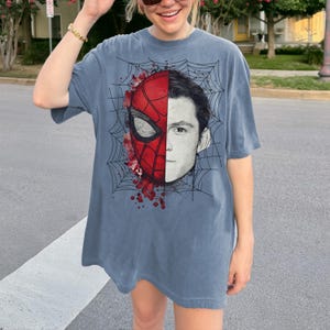 May include: A slate blue short-sleeve t-shirt featuring a graphic print of a split face. One side shows a red and black spider-themed mask with web patterns and red paint splatters, while the other side displays a black and white portrait of a human.