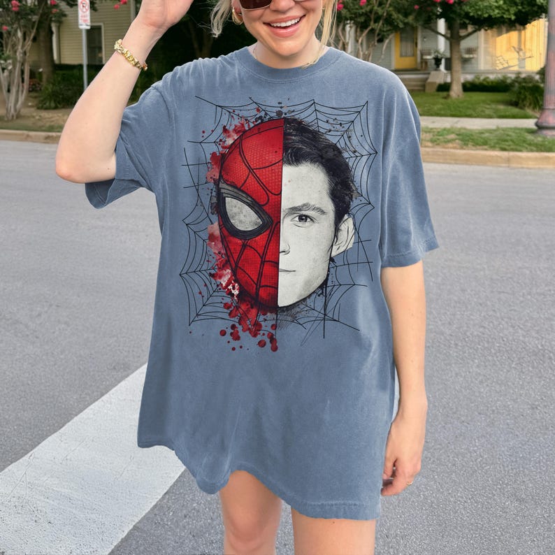 May include: A slate blue short-sleeve t-shirt featuring a graphic print of a split face. One side shows a red and black spider-themed mask with web patterns and red paint splatters, while the other side displays a black and white portrait of a human.