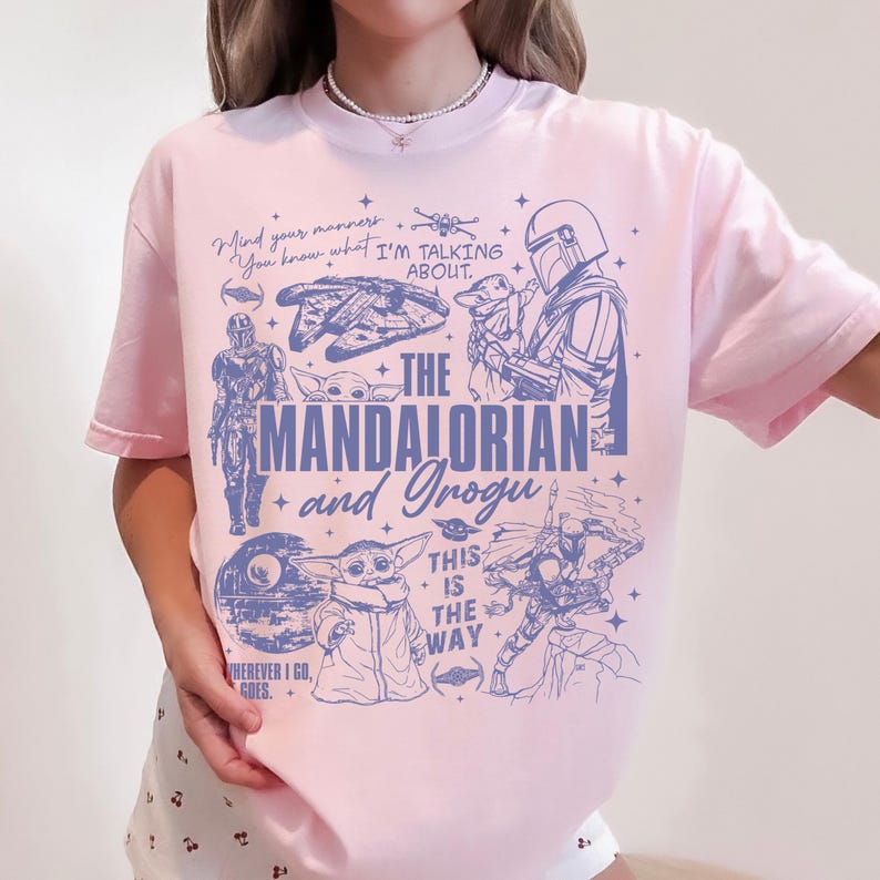 May include: A light pink t-shirt featuring a blue graphic print of The Mandalorian and Grogu. The design includes various character illustrations, star motifs, and text that reads: Mind your manners, I'm talking about, The Mandalorian and Grogu, This is the way.