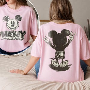 May include: A light pink short-sleeve t-shirt featuring a sketch-style graphic of Mickey Mouse on the front with the text MICKEY below it. The back of the shirt displays a large black graphic of Mickey Mouse from behind making hand gestures.