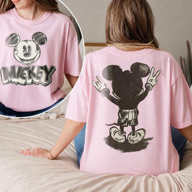 May include: A light pink short-sleeve t-shirt featuring a sketch-style graphic of Mickey Mouse on the front with the text MICKEY below it. The back of the shirt displays a large black graphic of Mickey Mouse from behind making hand gestures.