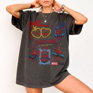 May include: A charcoal gray oversized t-shirt featuring multiple Spider-Man graphics, including heart-shaped and rectangular frames with red bows, spider web patterns, and the text Spider-Man Brand New Day and Marvel Studios printed in red and blue.