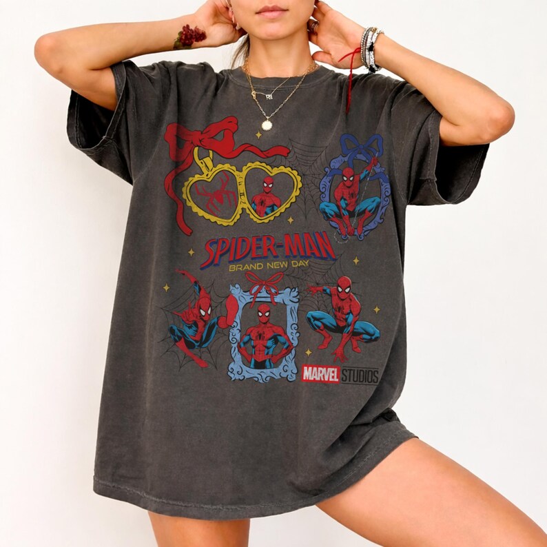 May include: A charcoal gray oversized t-shirt featuring multiple Spider-Man graphics, including heart-shaped and rectangular frames with red bows, spider web patterns, and the text Spider-Man Brand New Day and Marvel Studios printed in red and blue.