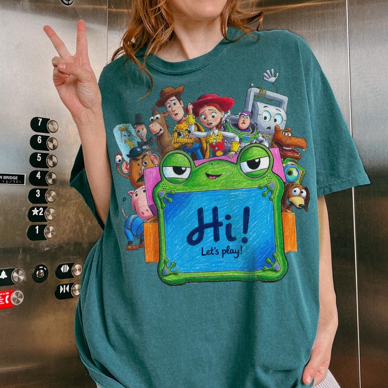 May include: A dark teal short-sleeve t-shirt featuring a graphic print of various animated characters surrounding a large green frog-shaped frame. The center of the frame is blue with the text "Hi! Let's play!" written in dark blue script.