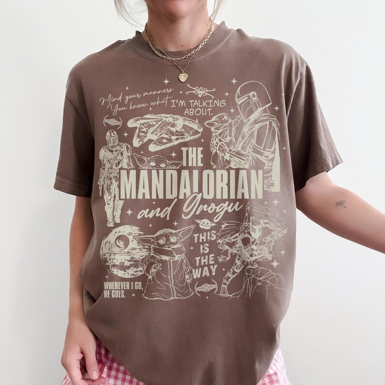May include: A brown short-sleeve t-shirt featuring a cream-colored graphic print of The Mandalorian and Grogu. The design includes line art of characters, the Millennium Falcon, the Death Star, and text reading The Mandalorian and Grogu.