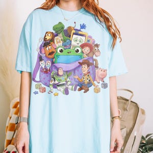 Op de afbeelding: Lichtblauw T-shirt met een kleurrijke afbeelding van personages uit de film "Toy Story". Het ontwerp bevat Woody, Buzz Lightyear en andere personages, samen met een paarse rugzak. Het shirt is een casual stijl met korte mouwen.