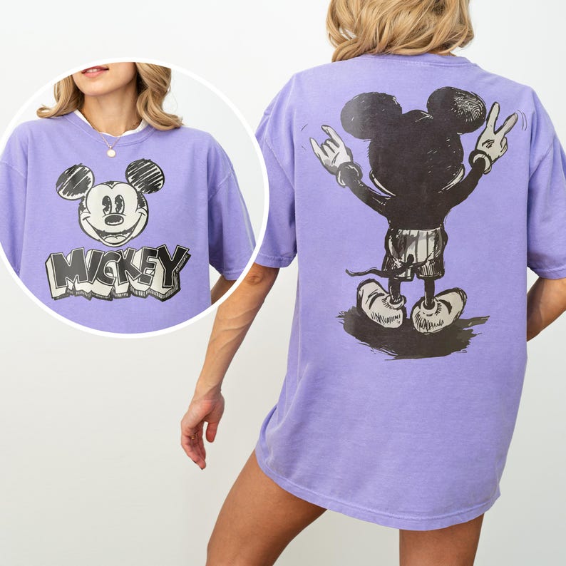 May include: A light purple t-shirt featuring a black and white sketch of Mickey Mouse on the front with the text "MICKEY" below. The back of the t-shirt displays a large black and white graphic of Mickey Mouse from behind, making a hand gesture.