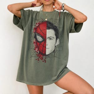May include: An olive green short-sleeve t-shirt featuring a split-face graphic print. One side displays a red and black Spider-Man mask, while the other side shows a black and white portrait of a young man, all set against a black spiderweb design.