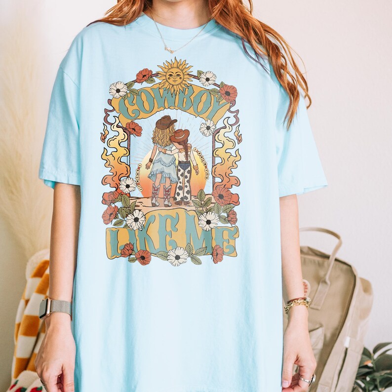 May include: A light blue t-shirt featuring a graphic print of two figures in cowboy hats standing with arms around each other. The design includes a sun, flowers, and the text "COWBOY LIKE ME" in a bold, retro-style font with orange and green tones.