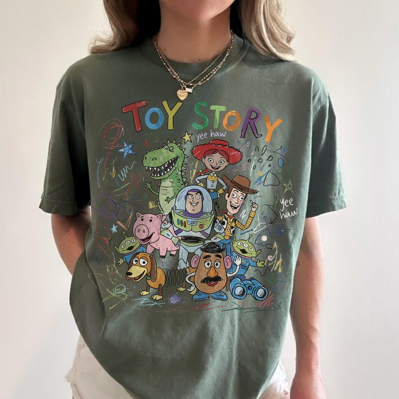 Peut inclure: T-shirt vert olive avec un motif color&eacute; des personnages du film "Toy Story". Le motif comprend les mots "Toy Story" en couleurs arc-en-ciel, ainsi que des dessins de Woody, Buzz l'&Eacute;clair et d'autres personnages.