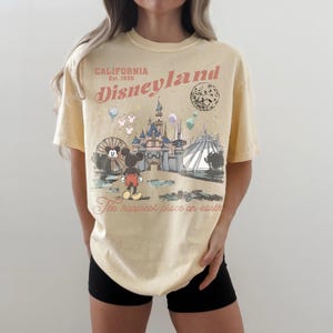 May include: A light yellow t-shirt featuring a Disneyland graphic. The design includes the text "California Est. 1955 Disneyland" and an illustration of Mickey Mouse, a castle, and other park attractions. The phrase "The happiest place on earth" is also printed on the shirt.
