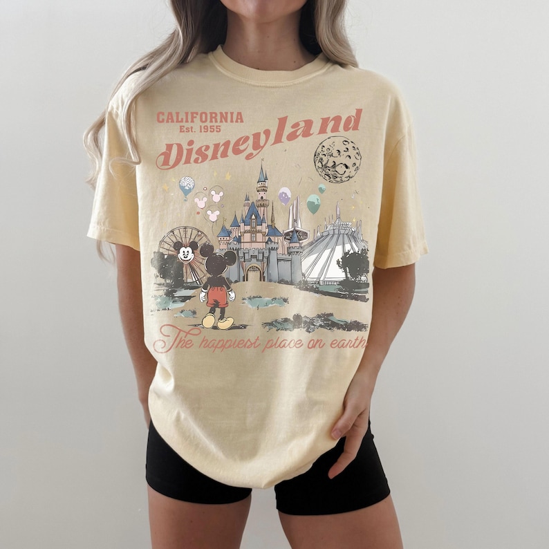 May include: A light yellow t-shirt featuring a Disneyland graphic. The design includes the text "California Est. 1955 Disneyland" and an illustration of Mickey Mouse, a castle, and other park attractions. The phrase "The happiest place on earth" is also printed on the shirt.