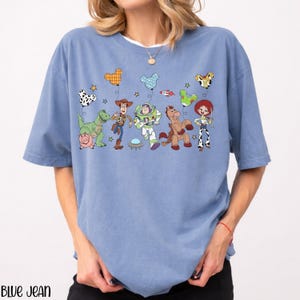 Puede incluir: Una camiseta azul de manga corta con un estampado gr&aacute;fico de personajes de Toy Story, incluyendo a Woody, Buzz Lightyear, Jessie, Rex, Perdig&oacute;n y Hamm. Los personajes sostienen globos coloridos con forma de orejas de Mickey Mouse.