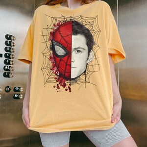 May include: A yellow short-sleeve t-shirt featuring a graphic print of a face split between a red Spider-Man mask and a realistic human portrait. The design includes black spiderweb line art and red paint splatter accents on the mask side.