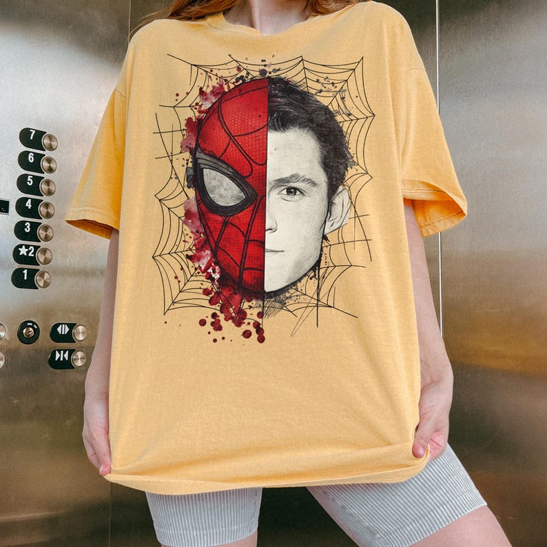 May include: A yellow short-sleeve t-shirt featuring a graphic print of a face split between a red Spider-Man mask and a realistic human portrait. The design includes black spiderweb line art and red paint splatter accents on the mask side.