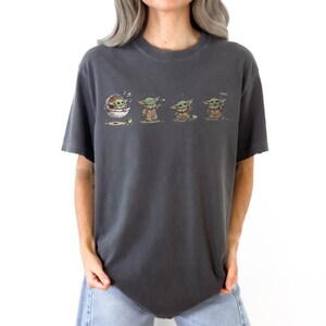 May include: A charcoal gray short-sleeve t-shirt featuring a graphic print of a small green creature in a series of four poses. The illustrations show the character in a floating pod, reaching for a frog, sitting with a cup, and standing.