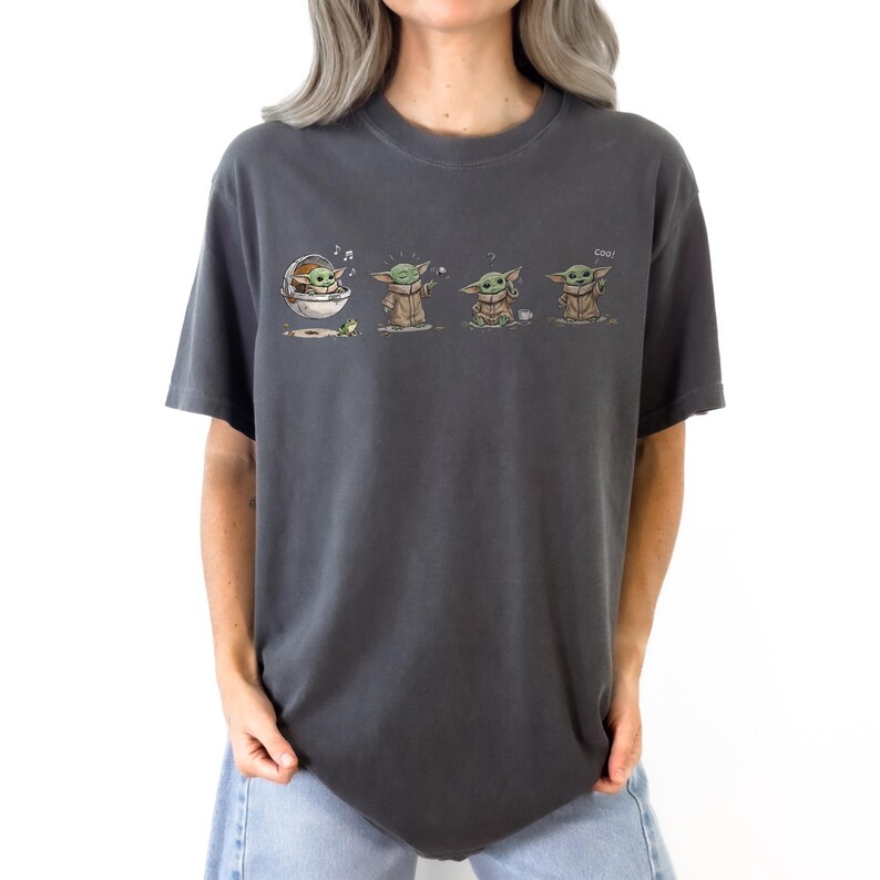 May include: A charcoal gray short-sleeve t-shirt featuring a graphic print of a small green creature in a series of four poses. The illustrations show the character in a floating pod, reaching for a frog, sitting with a cup, and standing.