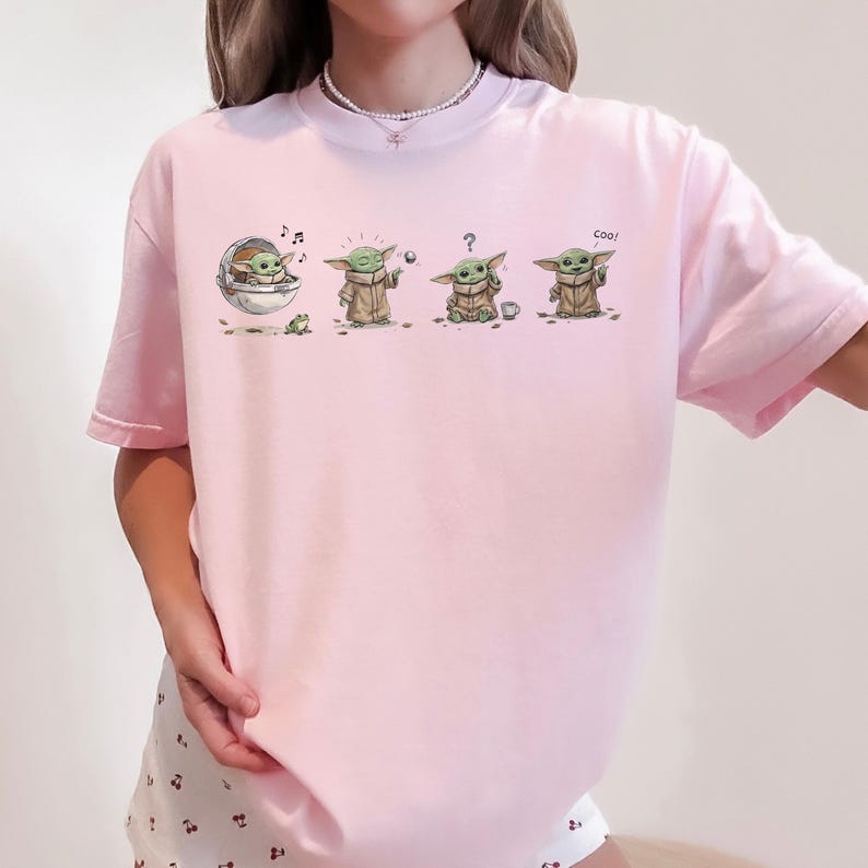 May include: A light pink t-shirt featuring a graphic print of a small green creature in four different poses. The illustrations show the character in a floating pod, using the force, looking confused with a question mark, and saying the text Coo!
