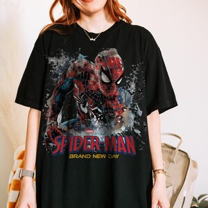 May include: A black short-sleeve t-shirt featuring a graphic print of Spider-Man in a red and blue suit. The design includes a paint splatter effect, the Marvel logo, and the text "SPIDER-MAN BRAND NEW DAY" in bold red and yellow lettering.