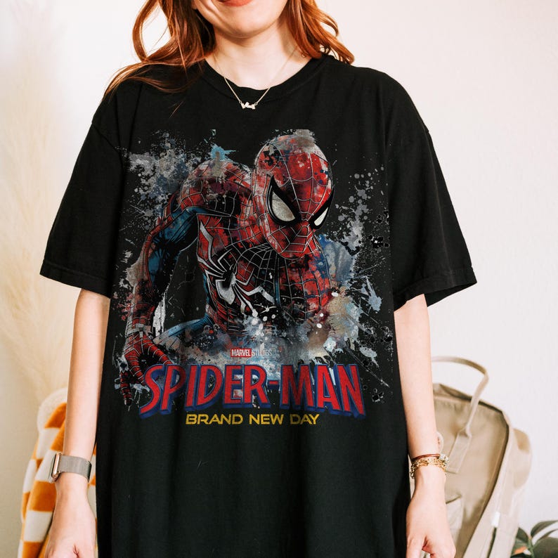 May include: A black short-sleeve t-shirt featuring a graphic print of Spider-Man in a red and blue suit. The design includes a paint splatter effect, the Marvel logo, and the text "SPIDER-MAN BRAND NEW DAY" in bold red and yellow lettering.